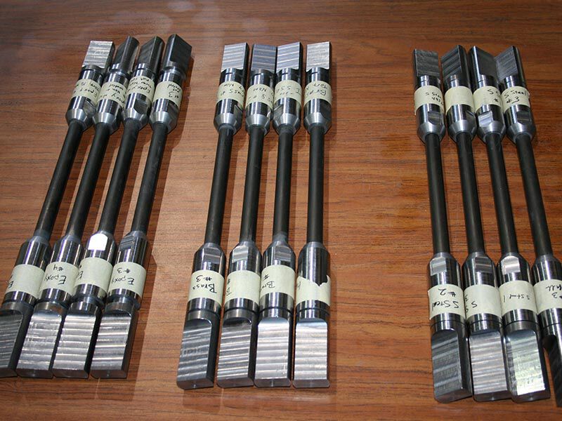 Row of Megalex™ sucker rods on a wooden desk. There are 12 rods in three groups of four. Each rod has a central carbon fiber column and metallic end-attachments.